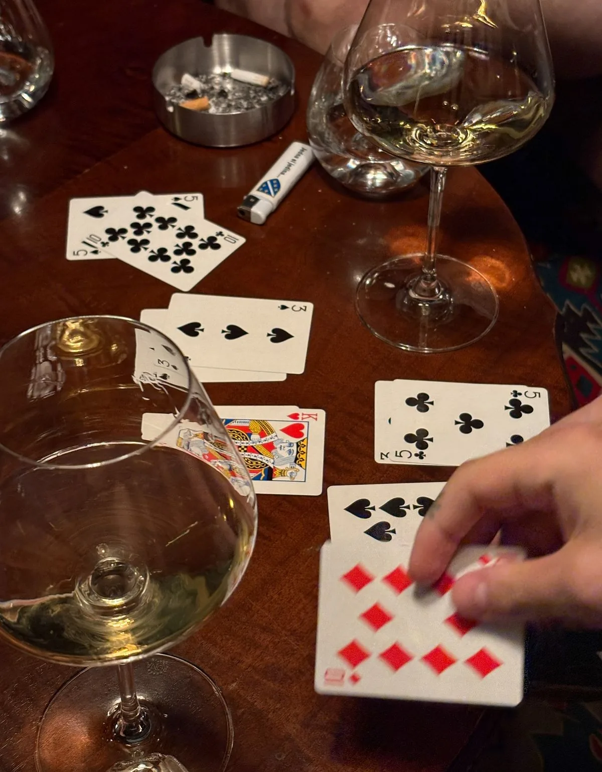 Cards in play on dark wood, a brass ashtray and two glasses of white wine.