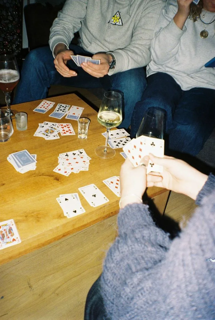 Friends on a couch playing cards over a wooden coffee table, sweaters and a glass of white wine.