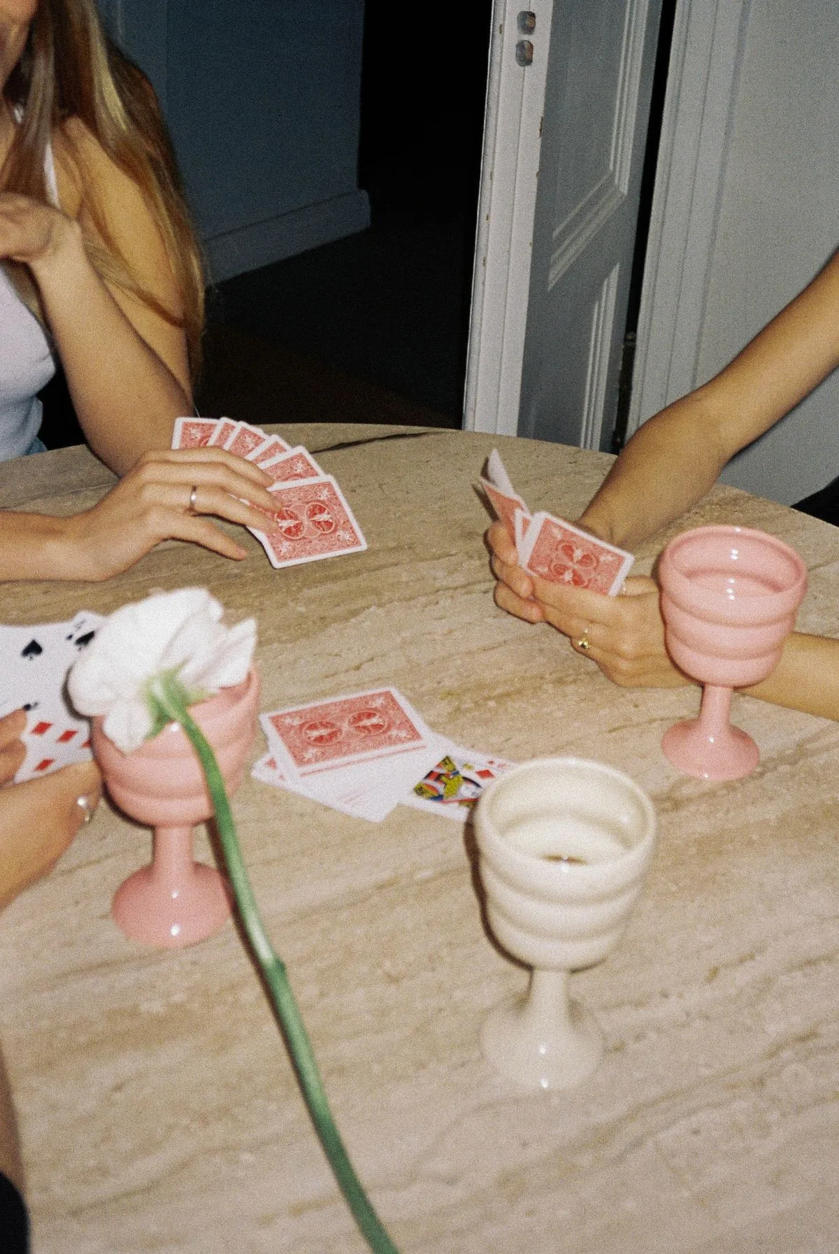 Friends around a stone table with pink and white goblets, fanned hands of cards mid-round.
