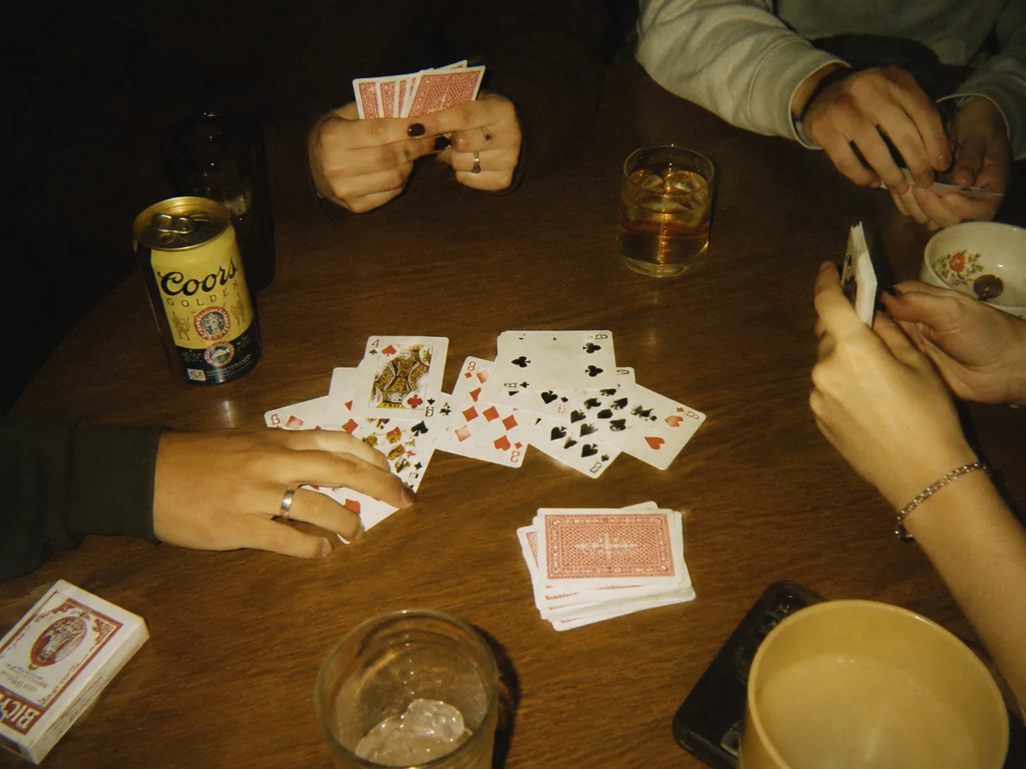 Four hands around a wooden table mid-game, a Coors can and a whiskey rocks glass alongside the scattered cards.