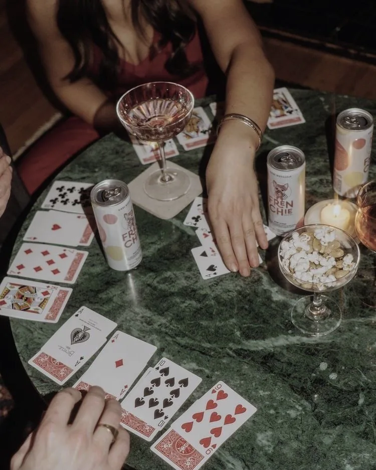 Cards spread across a green marble table, rosé in a coupe, friends mid-game.