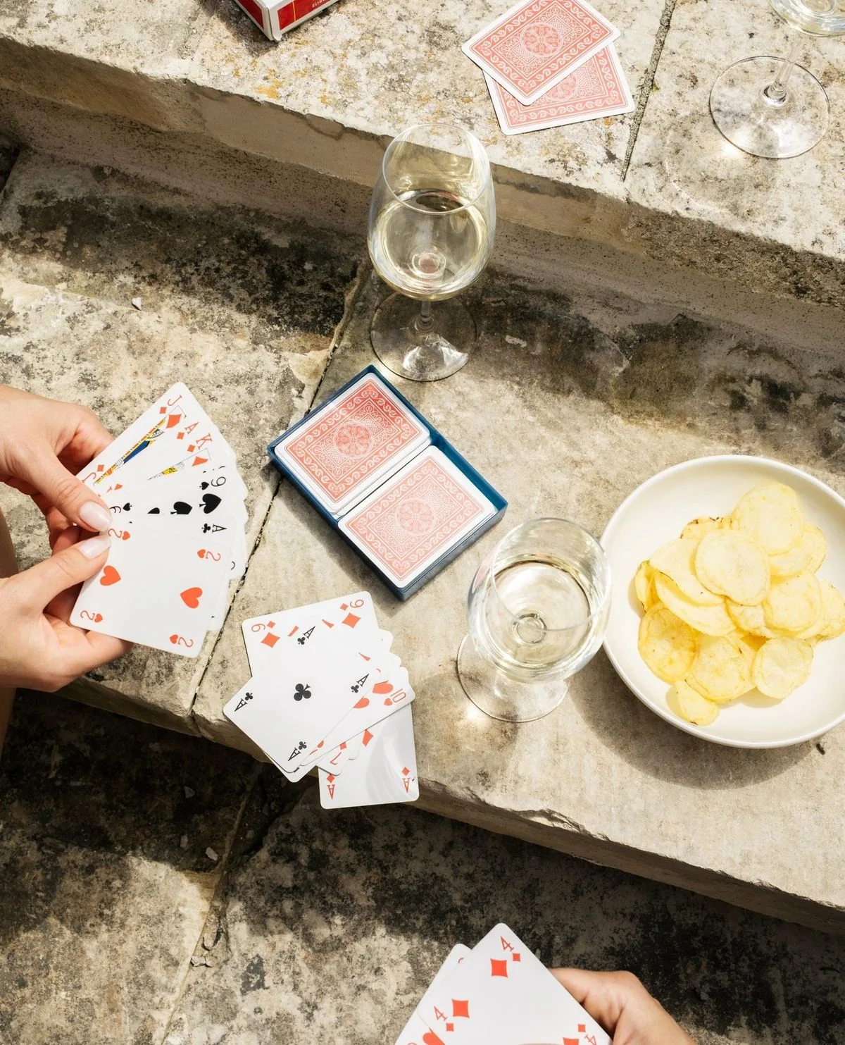 A hand of cards on weathered stone steps, two glasses of white wine and a bowl of crisps alongside.
